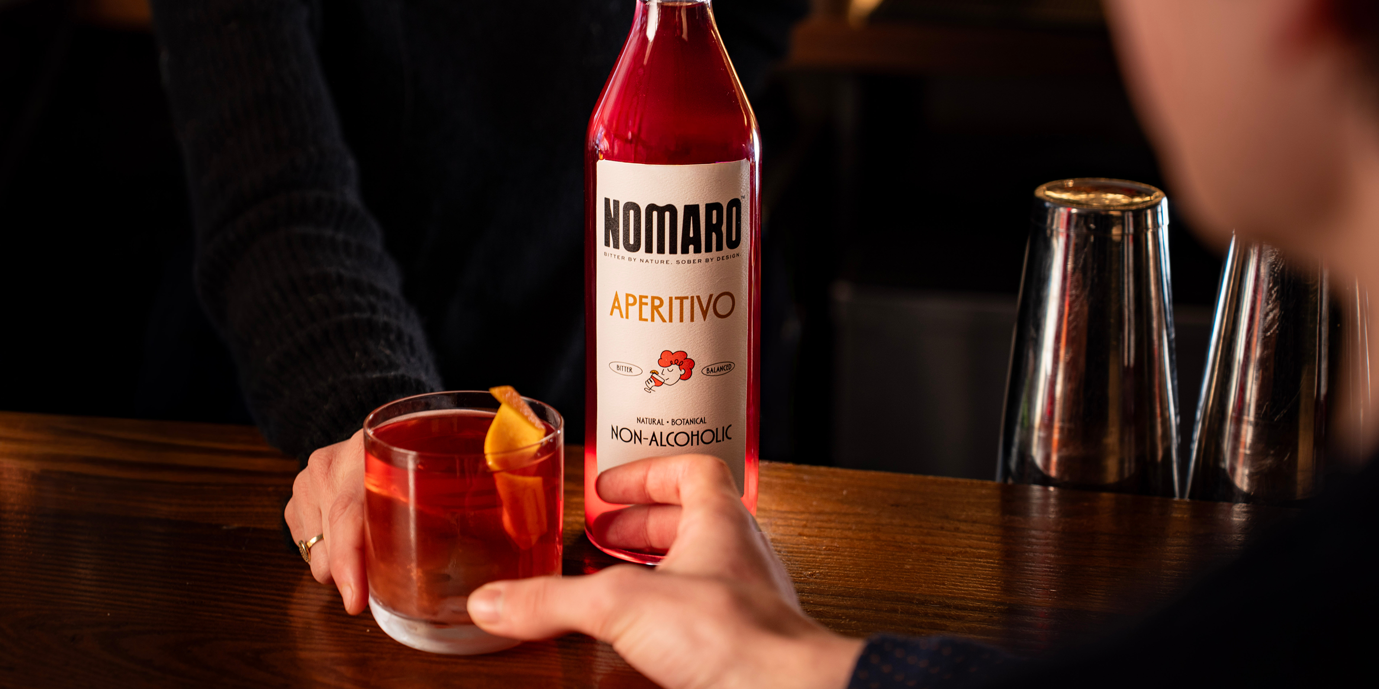 Bottle of Nomaro Aperitivo with a glass of a red drink on a bar counter.